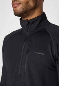 Zwarte fleece met rits en hoge kraag, voorzien van een textuurstof, een borstzak en een subtiele logo aan de voorkant.