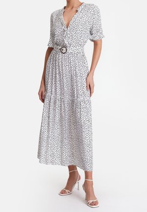 Vrouw draagt een witte midi-jurk met zwarte stippen, korte mouwen, een riem met een ronde gesp en witte sandalen met hoge hakken.