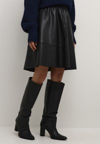 Jupe en cuir noir avec une taille élastique, coupe évasée, et bottes en cuir noir montantes jusqu'aux genoux, à bouts pointus et talons blocs.