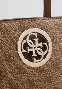 Tissu marron texturé pour sac à main avec motif en relief répété "GS" et un emblème logo rond en métal doré présentant des lettres entrelacées "G" et "S".