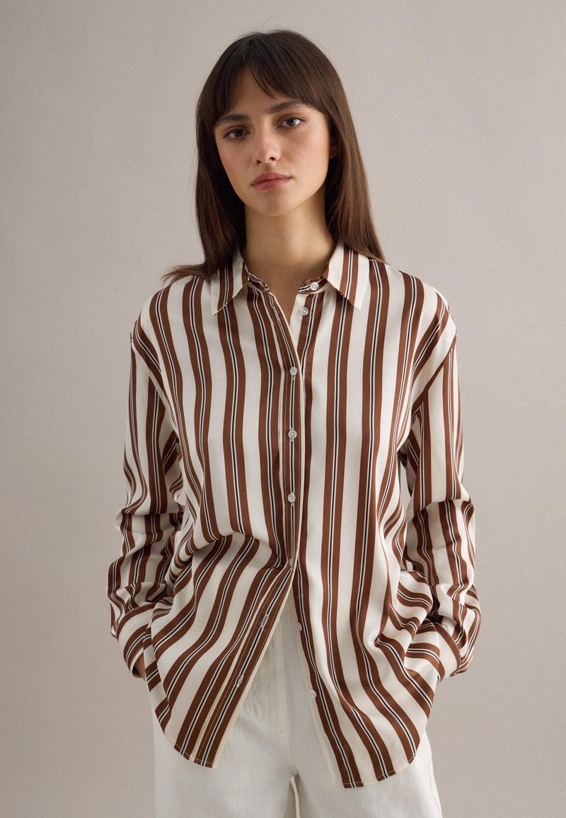 Mujer joven con cabello castaño lacio que lleva una camisa de botones con rayas verticales marrón y blanca, de pie con las manos en los bolsillos.