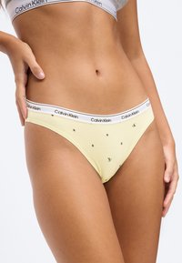 Žena v žltom spodnom prádle Calvin Klein s malými čiernymi hviezdičkami a bielym pásikom s logom, ruka položená na bok.