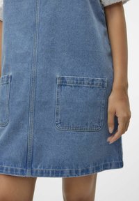 Denimklänning med stora framfickor, visande knähöjd och en persons hand som vilar bredvid klänningen.