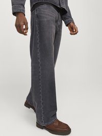 Mørk denim med vide ben og en falmet finish, kombinert med brune sko. Stoffet er robust; detaljer inkluderer søm langs sømmer og lommer.