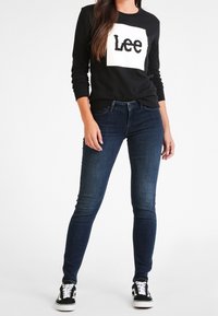 Kvinna iklädd en svart långärmad tröja med "Lee"-logotyp, mörkblå tajta jeans och svart-vita sneakers som står mot en vit bakgrund.