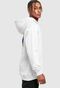 Sudadera blanca con un bolsillo canguro en la parte frontal, puños acanalados y dobladillo. Presenta un gráfico abstracto colorido en el lado izquierdo. La tela parece suave y texturizada.