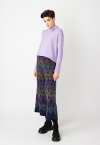 Violet megztinis su apykakle, derinamas su raštuotomis plačiomis kelnėmis, kurių dizainas sudarytas iš daugybės spalvotų geometrinių figūrų. Juodi tekstūruoti batai.