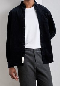 Camicia nera di velluto a coste con bottoni sopra una t-shirt bianca, abbinata a pantaloni grigi di velluto a coste, con taschino sul petto e dettagli di cuciture sottili.