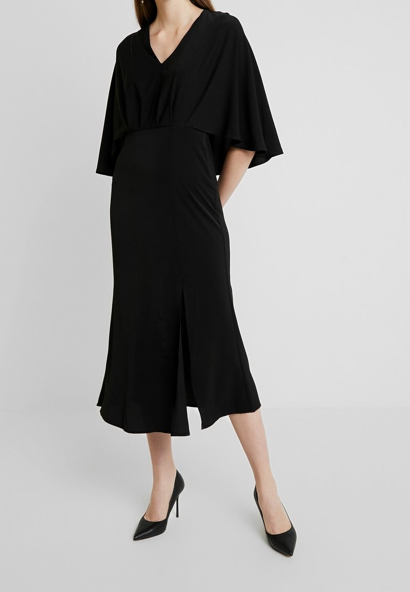 Femme portant une robe midi noire avec des manches larges et un décolleté en V, associée à des talons hauts noirs pointus, debout devant un fond uni.