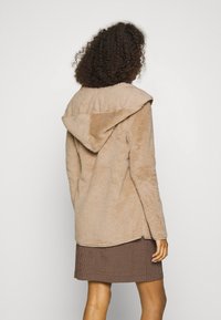 Tann fuzzy jacka med huva, som har en mjuk textur och en avslappnad passform. Bärs över en brun rutig kjol. Bakifrån visas en enkel design.