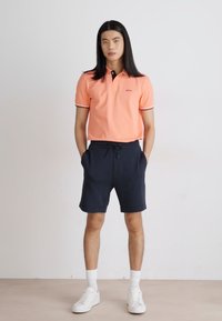 Oranžinė polo marškinė su kontrastinga juoda apykakle ir dryžuotais rankogaliais, derinama su mėlynais šortais ir baltais sportbačiais, stovinčiais ant neutralaus grindų paviršiaus.