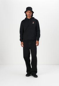 Felpa nera in cotone con tasca frontale e design di rosa ricamato; abbinata a pantaloni neri larghi e sneakers nere. Un cappello a secchiello completa il look.