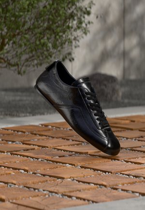 Schwarzer Ledersneaker mit Schnürsenkeln, schwebend über einem gemusterten Holz- und Steinboden, mit grünen Pflanzen und einer Betonwand im Hintergrund.