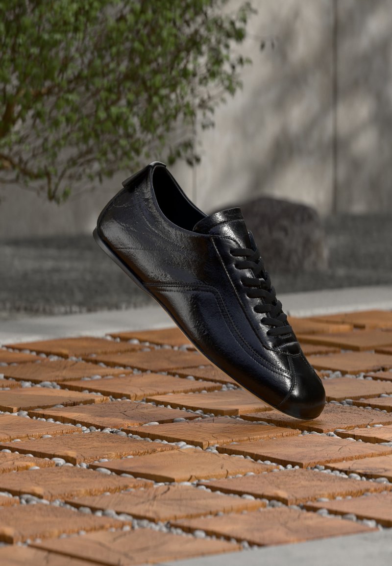 Μαύρο δερμάτινο αθλητικό παπούτσι με κορδόνια που αιωρείται πάνω από ένα διακοσμημένο ξύλινο και πέτρινο έδαφος, με πράσινα φυτά και έναν τσιμεντένιο τοίχο στο φόντο.