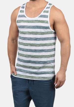 Mężczyzna w białym podkoszulku bez rękawów z niebiesko-zielonymi poziomymi paskami i ciemnoniebieskich spodniach, stojący z jedną ręką w kieszeni.