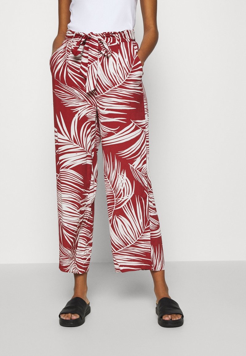 Pantalon rouge à jambes larges avec imprimé feuilles tropicales blanches, ceinture nouée à la taille, porté avec des sandales noires et un haut blanc sans manches.