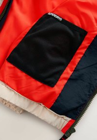 Interiér červenej a námorníckej bundy zobrazuje čiernu sieťovanú kapsu s labelom "SUPERDRY®" a béžový elastický pás blízko zeleného zipsu.