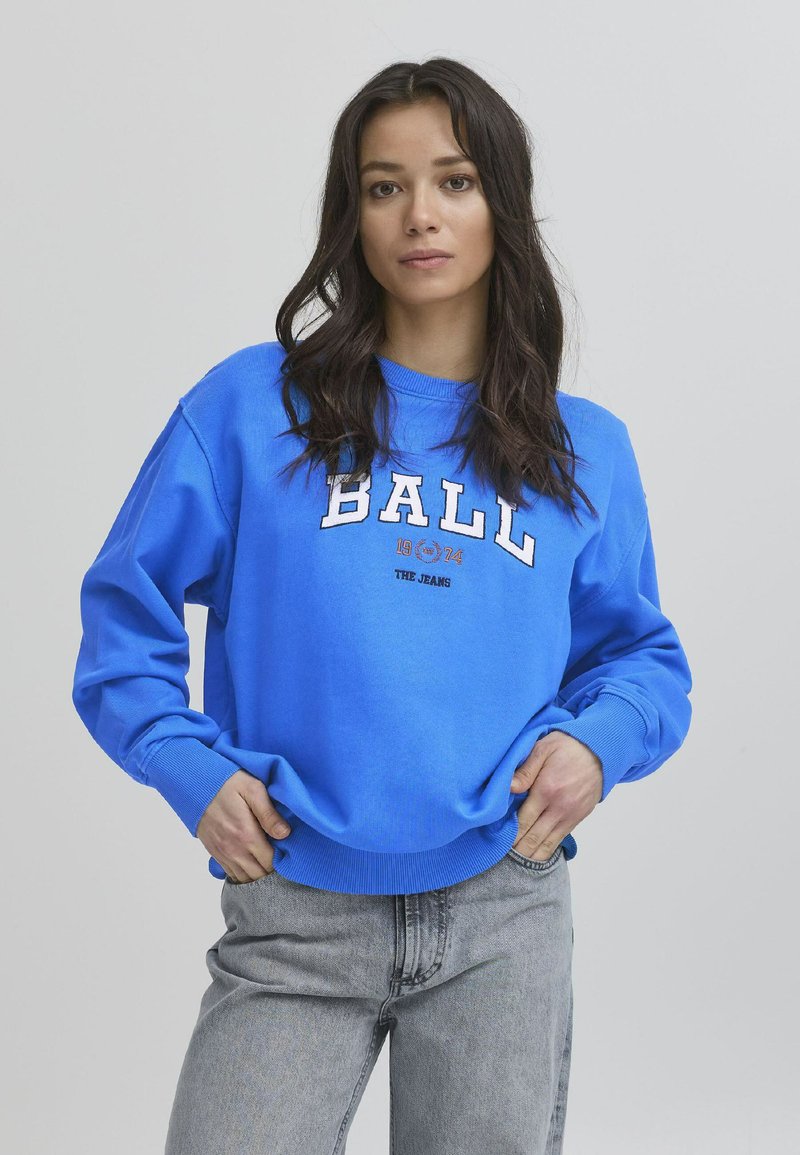 Blå hettegenser med lange ermer, ribbestrikkede mansjetter og hem. Har hvit og oransje tekst som sier "BALL 1904 THE JEANS." Brukt med lysegrå jeans.
