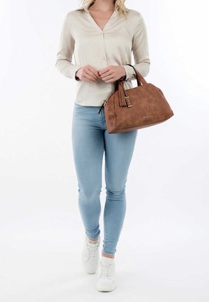 Vrouw draagt een beige blouse met lange mouwen, lichtblauwe strakke jeans, witte sneakers, en houdt een bruine suède handtas met franjes vast.