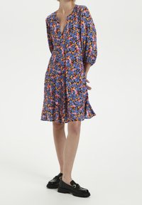 Robe florale avec un décolleté en V profond, des manches bouffantes et un ourlet évasé. Le matériau est léger et présente un mélange de teintes violettes, roses et oranges.