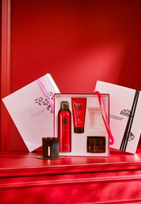 Röd presentlåda med ett svart ljus, två röda tuber och en burk i en vit låda med rosa band och blomsterprägling. Mot en röd bakgrund.