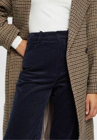 Brązowy i czarny płaszcz w deseń houndstooth noszony na białej koszuli, w parze z granatowymi spodniami z kordurowego materiału o teksturowym wykończeniu i z przodu zapinanymi na guzik.