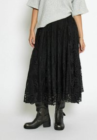 Gonna maxi in pizzo nero con motivi intricati, design stratificato e finitura testurizzata. Abbinata a stivali neri alla caviglia per un look completo.