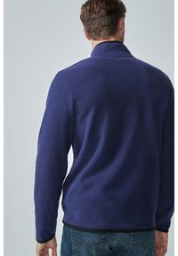 Mann trägt eine dunkelblaue Fleecejacke und blaue Jeans, steht mit dem Rücken zur Kamera vor einem schlichten hellen Hintergrund.