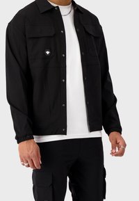 Czarna casualowa kurtka zapinana na guziki, z dwoma kieszeniami na piersi i naszywką z logo. Posiada luźny fason i ściągacze na mankietach. Nosi się ją na białą koszulkę.