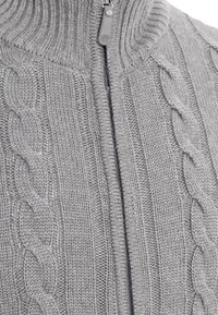 Gros plan d'un pull gris en tricot avec un motif torsadé et une fermeture éclair centrale partiellement fermée.