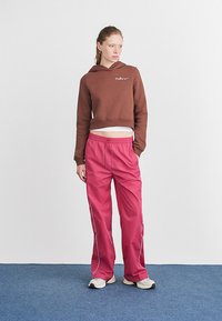 Καφέ κοντό hoodie με λευκό λογότυπο Nike, συνδυασμένο με χαλαρούς ροζ παντελόνες με λευκές λεπτομέρειες και ελαστική μέση. Λευκά αθλητικά παπούτσια.