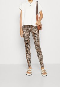 Witte kurzemouwen top met een kraag, lichtbruine legging met luipaardprint, en witte sandalen met een decoratieve bloemenaccent. Bruine schoudertas.