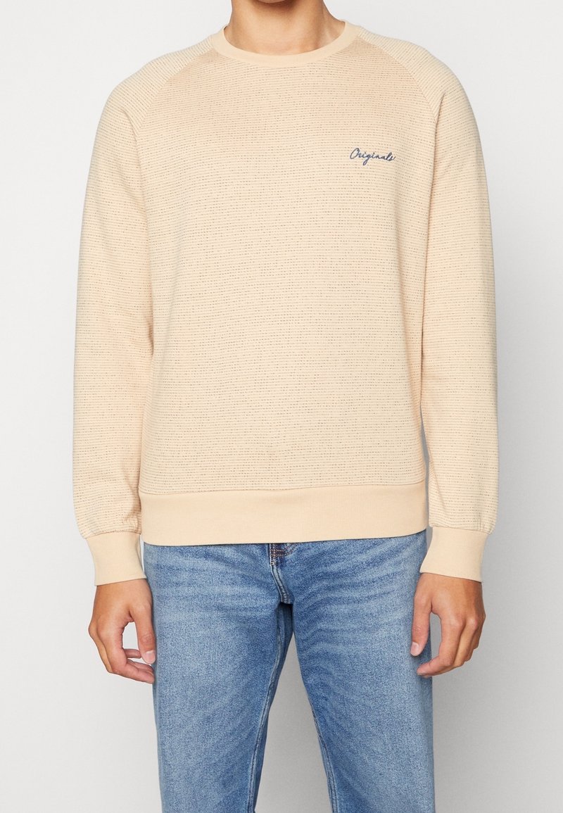 Pull beige texturé à manches longues avec poignets et ourlet côtelés, petit texte « Originals » sur la poitrine gauche, porté avec un jean bleu.
