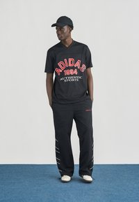 Άνδρας που φοράει μαύρα αθλητικά ρούχα Adidas με κόκκινο και άσπρο λογότυπο, μαύρο καπέλο και άσπρα παπούτσια, στέκεται σε μπλε χαλί μπροστά σε γκρίζο τοίχο.