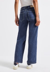 Jean en denim �à jambes larges dans un denim bleu foncé, avec une taille structurée, des poches arrière et une texture légèrement délavée. Porté avec des baskets blanches.