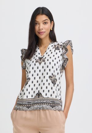 Femme aux longs cheveux foncés portant un chemisier sans manches blanc à motifs avec des épaules volantées et un pantalon beige, debout devant un fond uni.