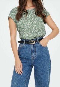 Top verde con stampa floreale e maniche corte, abbinato a jeans in denim blu a vita alta e una cintura nera con una fibbia dorata.
