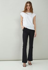 Woman standing with hand in pocket wearing white short-sleeve top, black pants, and black open-toe heels against a neutral background.