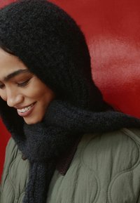 Schwarze gestrickte Kapuze und Schal aus weichem Material, kombiniert mit einer gesteppten grünen Jacke mit kreisförmigen Mustern. Hintergrund in leuchtendem Rot.