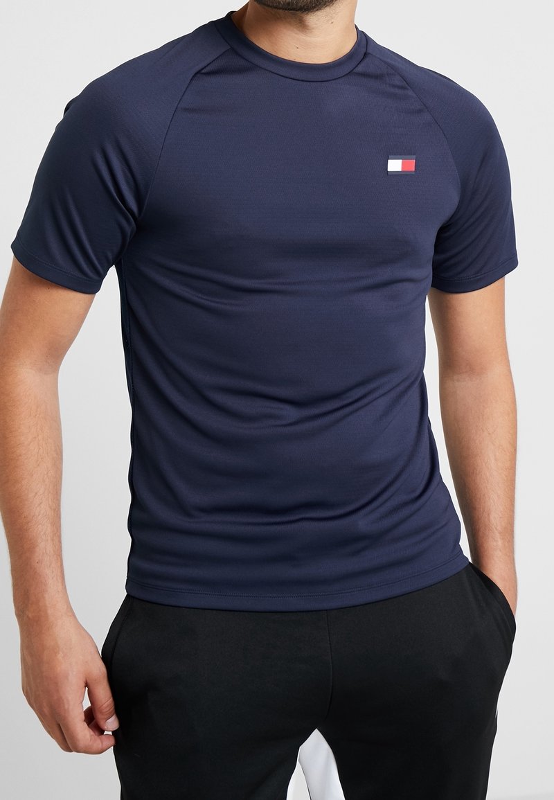 Homme portant un t-shirt de sport ajusté bleu marine à manches courtes avec un petit logo rectangulaire rouge, blanc et bleu sur la poitrine.