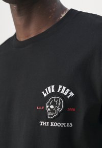 Czarna bawełniana koszulka z grafiką czaszki oraz napisem "LIVE FAST" i "THE KOOPLES" haftowanym na biało i czerwono. Klasyczny krój z okrągłym dekoltem.