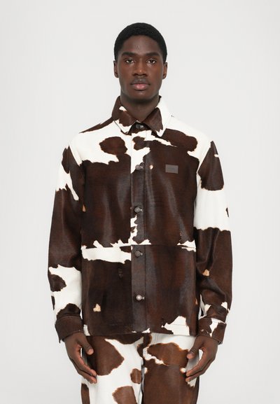 Veste chemise à motif vache marron et blanc en tissu texturé, dotée d'un col, de boutons-pression et d'un patch logo discret sur la poitrine.