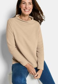 Maglietta beige a maniche lunghe a coste con collo a cascata, indossata con jeans blu. Tessuto liscio e vestibilità comoda, che mette in risalto uno stile informale.