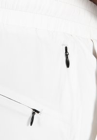 Pantalones cortos de tela blanca con cintura elástica fruncida y bolsillos con cremallera negra, que destacan por su textura suave y diseño minimalista.