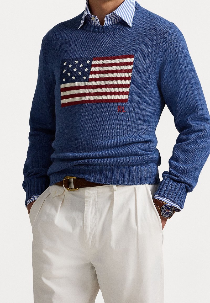 Homme portant un pull bleu avec motif du drapeau américain, pantalon blanc, chemise à col rayé, ceinture marron et montre-bracelet.