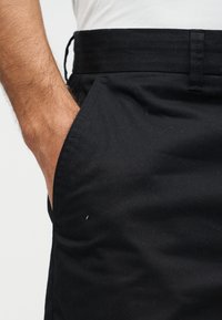 La main d'un homme reposant dans la poche avant d'un pantalon noir, portant une chemise blanche rentrée dans la ceinture.