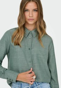 Camisa verde con botones, tejido texturizado, cuello clásico y mangas largas con botones en los puños. Se lleva con vaqueros azul claro.