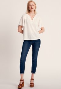 Top bianco a maniche corte con scollatura a V. Abbinato a jeans attillati blu scuro e sandali con zeppa marroni, dotati di cinturini regolabili.