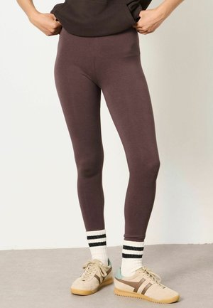 Person wearing brown leggings, white socks with black stripes, beige sneakers, and a dark top with hands pulling the top up slightly.
