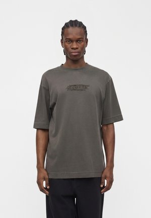Ung mand med flettet hår iført en løs mørkegrå T-shirt med et svagt logo i midten og sorte bukser, stående foran en ensfarvet baggrund.